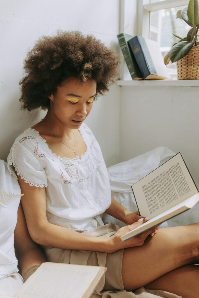 pexels-photo-5084674-5084674 A young woman with natural hair enjoys reading a book by the window with warm sunlight.